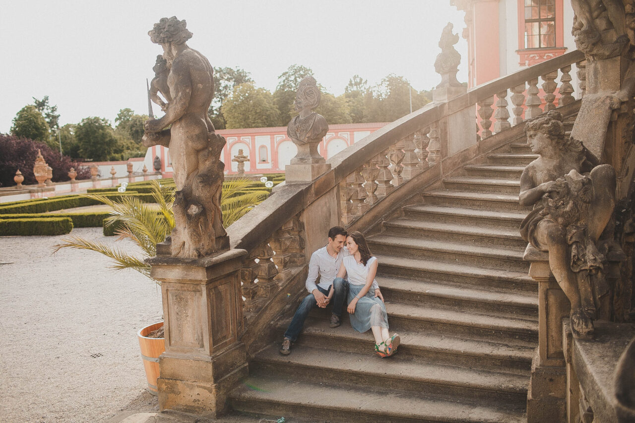 una sesión de fotos de una enamorade pareja frente a un castillo grande y hermoso. Se sientan en las escaleras y se miran tomados de la mano.